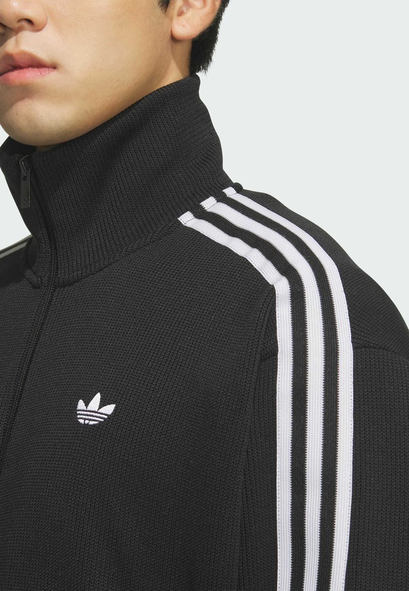 Man draagt een zwarte gebreide jas met hoge kraag met wit Adidas trefoil logo en drie witte strepen op de schouder en mouw.