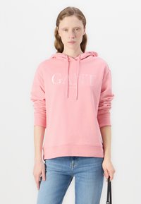 Rosa hoodie i mjukt tyg, med en framsidan logo i silver. Den har dragsko i huvan och ribbade muddar, tillsammans med ljusblå jeans.