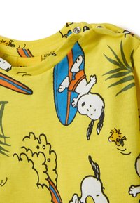 Camicetta gialla a maniche corte con personaggi dei cartoni animati e tavole da surf, caratterizzata da una consistenza morbida e chiusura a scatto sul colletto.