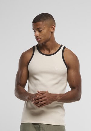 Svalnatý muž s krátkymi vlasmi, oblečený v béžovom tielku s čiernym lemom, pozerá sa nadol, ruky zložené pred sebou na jednoliatom pozadí.