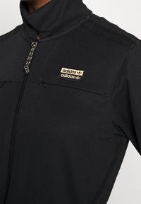 Černá sportovní bunda s vysokým límcem, zipovým zapínáním, texturovanou látkou a zlatým logem Adidas na prsou. Obsahuje dvě kapsy na zip.