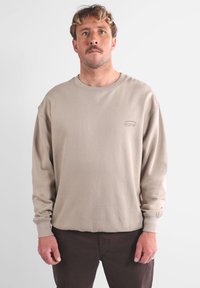 Sweatshirt beige à manches longues en mélange de coton doux, avec un col rond et des manches à poignets, affichant un petit logo sur la poitrine.