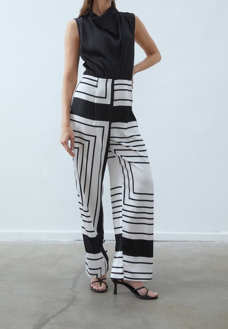 Femme portant un haut noir sans manches et un pantalon large taille haute avec des motifs géométriques noirs et blancs, debout sur un sol en béton.