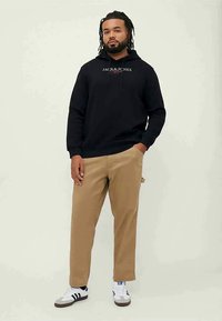Svart hoodie med vit logotyp på bröstet, i kombination med ljusbruna byxor och vita sneakers med svarta ränder. Enkl bakgrund, stående ställning.