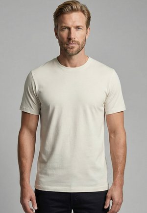 Blonde man met lichte baard draagt een effen gebroken wit T-shirt met korte mouwen en zwarte broek, staand tegen een grijze achtergrond.