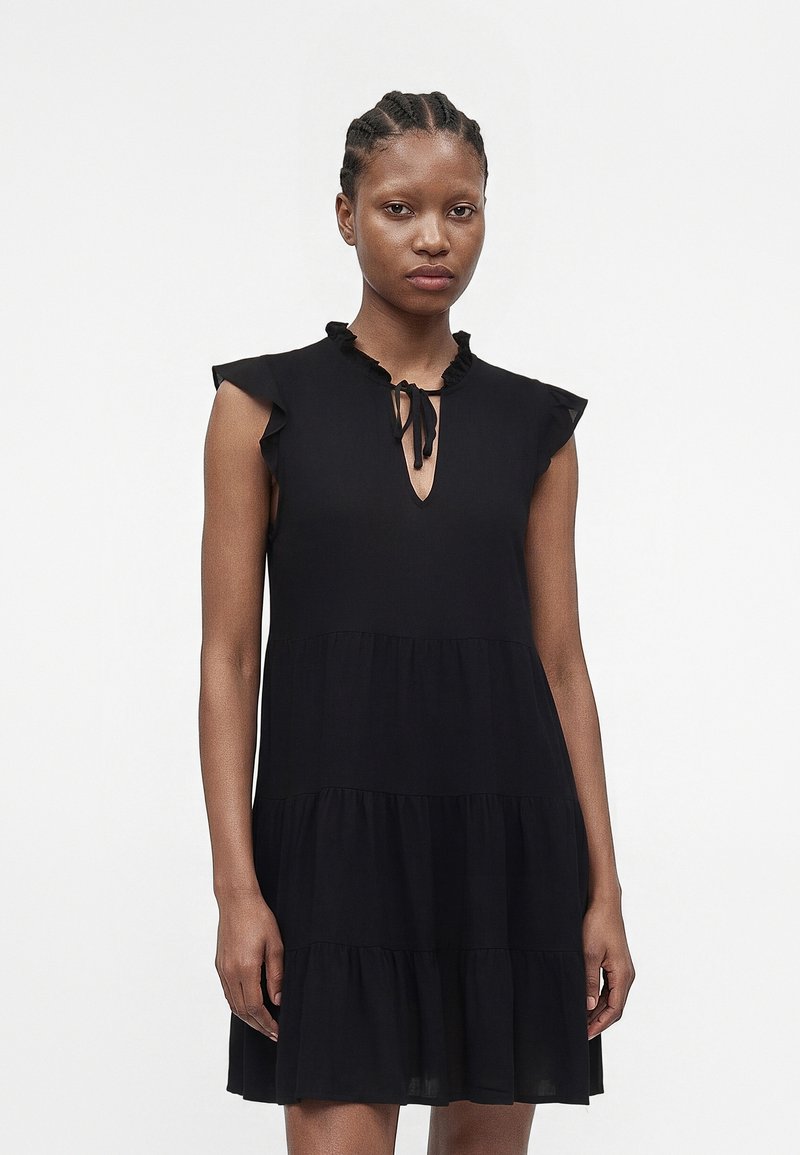 Γυναίκα με πλεξούδες που φοράει αμάνικο μαύρο φόρεμα με στρώσεις, με βολάν στον γιακά και κορδόνι στο λαιμό, που στέκεται μπροστά σε λευκό φόντο.