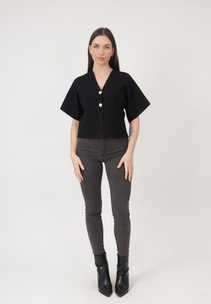 Ung kvinde med langt mørkt hår iført en sort knaplukket top, mørkegrå skinny jeans og sorte ankelstøvler, stående mod en ensfarvet baggrund.