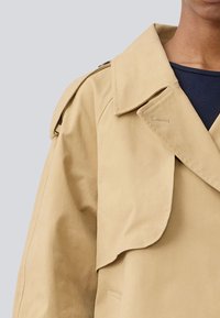Beige trenchcoat med store revers og en blød tekstur. Har en enkelt knaplukning og en sidelomme, hvilket skaber en afslappet pasform.