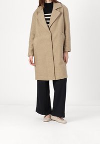 Béžový, texturovaný kabát s klopovým límcem, bočními kapsami a volným střihem, nošený přes černé kalhoty s širokými nohavicemi a světle zbarvenou obuv.