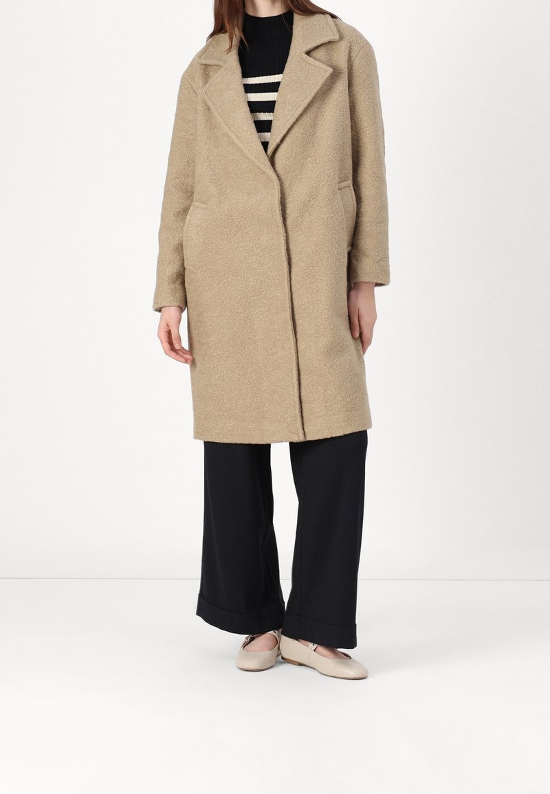 Béžový, texturovaný kabát s klopovým límcem, bočními kapsami a volným střihem, nošený přes černé kalhoty s širokými nohavicemi a světle zbarvenou obuv.