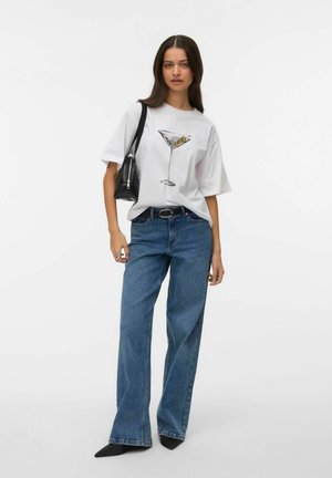 Ung kvinde iført en hvid T-shirt med tryk af en martiniglas, blå viddebenede jeans, sort bælte, sort håndtaske og sorte spidse sko.