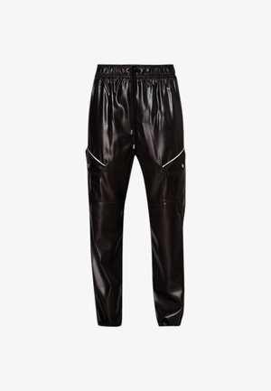 Kobieta ubrana w białą koszulę z czarnym obszyciem i guzikami, z paskiem w talii, czarne spodnie ze sztucznej skóry z kieszeniami, czarne szpilki i trzyma czarną łańcuszkową torebkę.