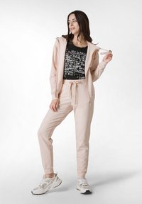 Chándal rosa claro con mangas largas y capucha, que incluye pantalones ajustados. Camiseta gráfica negra con texto blanco debajo. Zapatillas blancas.