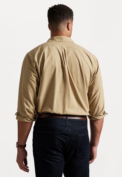 Khaki gombos ing feltekert ujjal, hátsó pliszével és sima textúrával, sötét farmernadrággal és barna övvel párosítva.