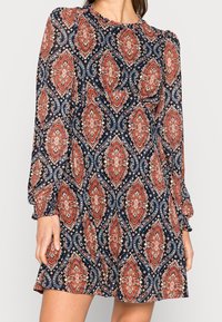 Mörkblå långärmad klänning med ett paisleymönster i rött, vitt och krämfärgat. Flödig tyg med veckade detaljer vid halsringningen och ärmarna.