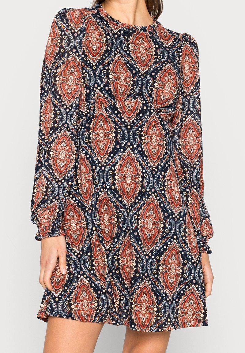 Mörkblå långärmad klänning med ett paisleymönster i rött, vitt och krämfärgat. Flödig tyg med veckade detaljer vid halsringningen och ärmarna.