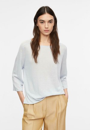 Jeune femme aux longs cheveux bruns portant un pull ample gris clair et un pantalon plissé beige, les mains dans les poches, debout sur fond blanc.