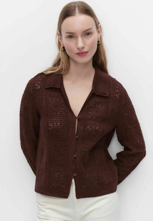 Kobieta o prostych blond włosach nosi ciemnobrązowy sweter na guziki na szydełku i białe spodnie, stojąc na tle jednolitego białego tła.