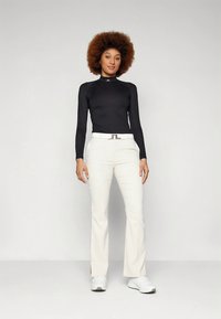 Kvinna med afrofrisyr som bär svart långärmad topp, vitt bälte, vita byxor och vita sneakers, stående mot en enfärgad ljus bakgrund.