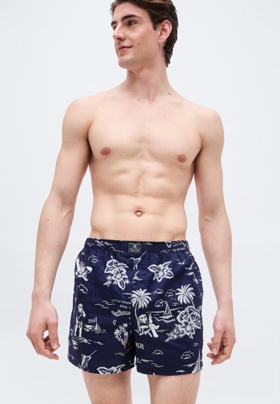 Άνδρας χωρίς μπλούζα που φοράει μπλε ναυτικό μαγιό με λευκά μοτίβα τροπικά και θεματικά ιστιοπλοΐας, στέκεται μπροστά σε απλό ανοιχτό φόντο.