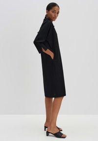 Schwarzes, knielanges Kleid mit Dreiviertelärmeln, Seitentaschen und lockerer Passform. Glatte Textur mit minimalistischem Design. Trägt man zu schwarzen Rutschen-Sandalen.