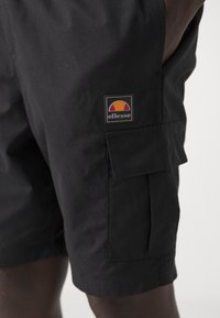 Black cargo shorts with textured fabric, featuring a side pocket, and an ellesse logo patch with a colored arc design.