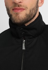 Veste noire avec col montant, dotée d'une fermeture éclair devant et d'une fermeture à boutons-pression. Texture de tissu lisse avec un éclat subtil.