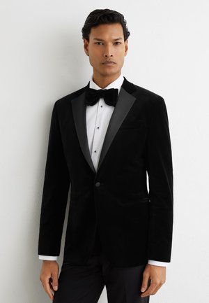 Homme portant une veste de smoking en velours noir avec des revers en satin, une chemise blanche et un nœud papillon noir, debout contre un mur blanc.