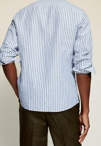 Striped shirt in light blue and white cotton, featuring rolled sleeves and a curved hem, viewed from the back. Linen pants visible below.