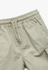 Pantalones cortos de un tono caqui claro, fabricados con un tejido suave, cuentan con una cinturilla elástica con cordón, bolsillos laterales y un bolsillo tipo cargo.