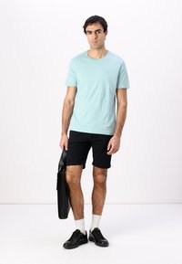 Világoskék pamut póló, fekete rövidnadrág, fekete sportcipő, fehér zokni, egy fekete táskát tart. A modell egy sima háttér előtt áll.