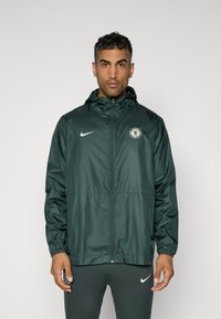 Dark green Nike windbreaker jacket with a hood, featuring a zip front, elastic cuffs, and a Chelsea FC badge on the left chest.
