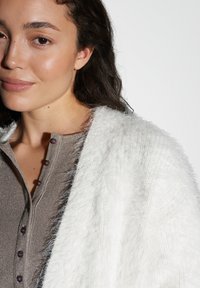 Jeune femme aux longs cheveux bruns portant un cardigan en peluche blanc sur un haut marron à côtes boutonné, devant un fond blanc uni.