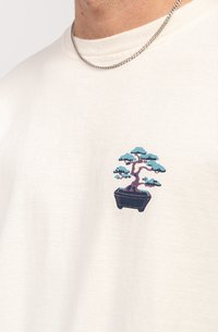 Krēmveida kokvilnas T-krekls ar nelielu zilo un purpura krāsā izšūtu bonsai koku kreisajā krūtīs. Vienkāršs, noapaļota kakla dizains.