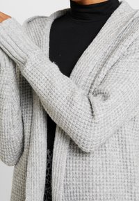 Gray knit cardigan with a textured pattern, open front, and ribbed cuffs. Worn over a black turtleneck, showcasing layered fabric details.