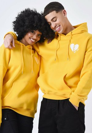 Las sudaderas con capucha amarillas presentan bolsillos frontales, cordones y un gráfico de un corazón roto. Se observa un tejido suave con un corte relajado.