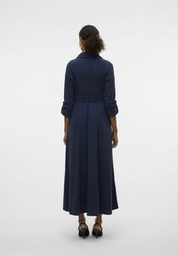Robe longue bleu marine avec un col, taille ceinturée et manches longues retroussées. Tissu doux avec un design fluide, atteignant les chevilles.