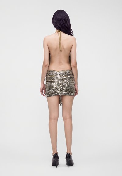 Femme aux longs cheveux foncés portant une mini-robe dos nu en sequins dorés et des talons hauts noirs, debout de dos devant un fond uni.