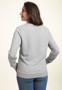 Mujer con cabello castaño que lleva puesta una sudadera gris clara de manga larga simple y jeans azules, de pie con las manos en las caderas, mirando hacia la izquierda.