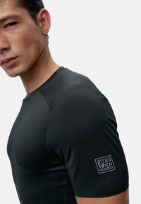 Melns īsām piedurknēm sporta krekls, izgatavots no gludas, stiepjamas auduma; ar kvadrātveida logo uzlīmi uz kreisās piedurknes.