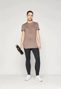 Helepruun lühikeste varrukatega topp, mustad retuusid, hoides musta veepudelit. Tossud on valged, taust on neutraalne. Pehme, sile kangatekstuur.