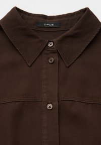 Camicia marrone scuro con collo e tre bottoni visibili, con un'etichetta nera con la scritta "OPUS" cucita all'interno del colletto.