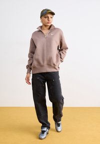 Felpa leggera marrone chiaro con zip a quarti e logo Nike, abbinata a jeans sformati scuri con due tasche frontali e scarpe da ginnastica grigie.