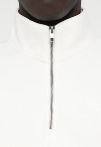 Balta jaukta kakla pūka ar gludu tekstūru, kas aprīkota ar sudraba metāla rāvējslēdzēju un augstu apkakli, lai nodrošinātu papildu siltumu.