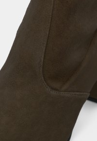 Bottine en suède marron avec une texture lisse, une forme arrondie et des coutures latérales. Présente un talon noir solide pour la stabilité.