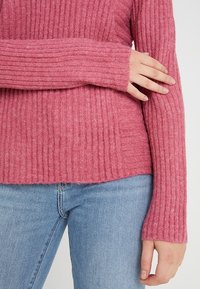 Pink ribbed sweater with long sleeves, soft texture, and a relaxed fit, paired with light blue jeans. Features a hem that sits below the waist.