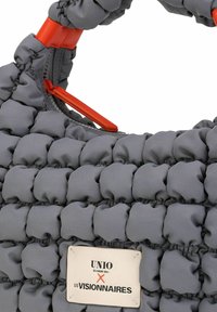 Grijze gewatteerde handtas met een getextureerd ontwerp, voorzien van opvallende rode accenten en een label met de tekst "UNIO HAMBURG X LES VISIONNAIRES."