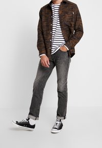 Chemise à boutons marron à motifs portée sur un t-shirt rayé blanc et marine, jeans gris et baskets noires. Tenue décontractée avec des textures contrastées.