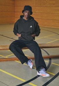 Hanorac negru Adidas, blugi negri cu model de frunze, adidași gri și portocalii, așezat pe o bancă de lemn într-o sală de sport cu linii de exerciții.