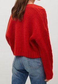 Pull rouge en maille avec une coupe courte, manches longues texturées avec des motifs en torsades, et un ourlet côtelé, porté avec un jean bleu.